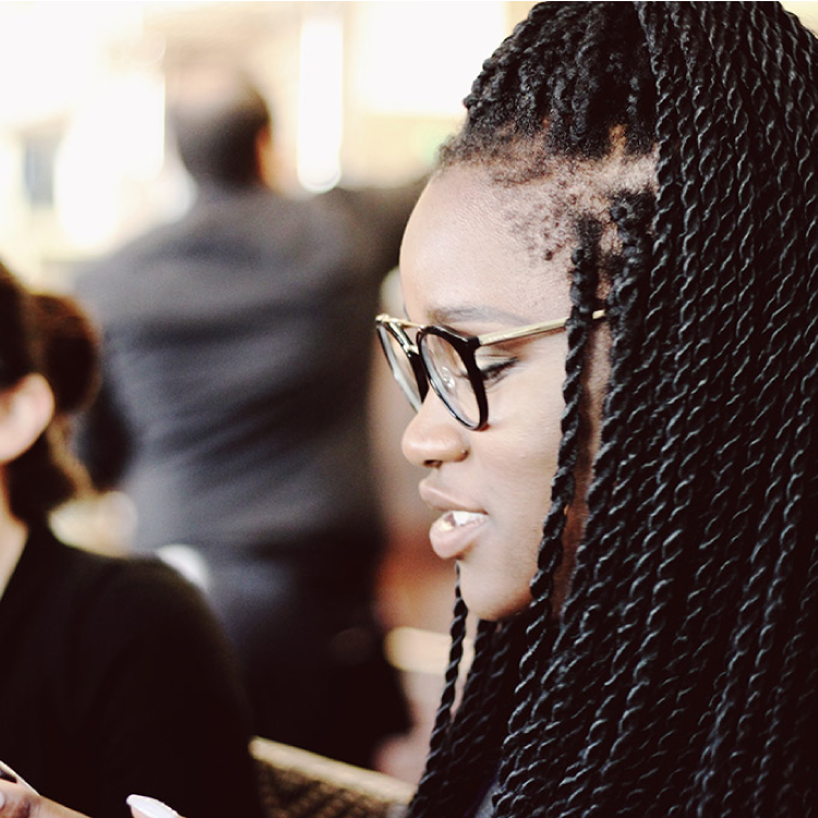 A young woman with long braided hair and glasses engaged in a conversation in a professional setting, with blurred figures in the background.