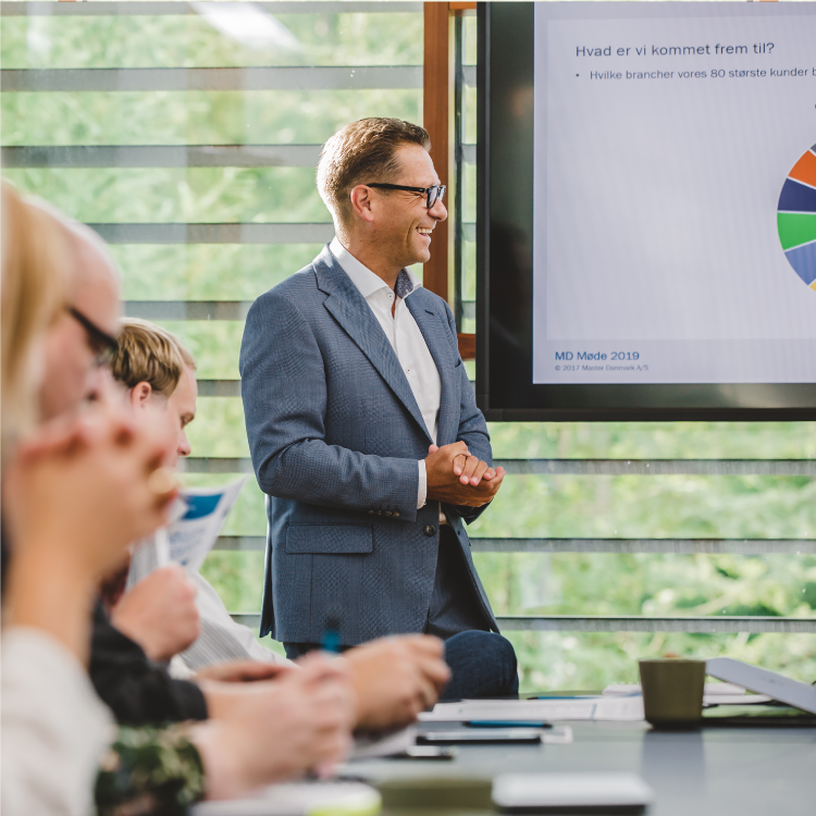A man in a blue suit and glasses stands smiling in front of a presentation screen displaying a pie chart. Seated attendees listen attentively, holding documents and taking notes.
